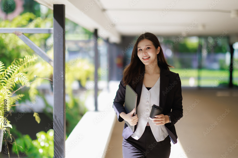 Fototapeta premium Asian professional businesswoman confidently walking outdoor through a modern office building hallway. Carrying a laptop and a coffee cup. Embodying success. Career advancement