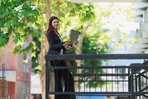 Young Asian businesswoman smiling, holding a laptop and coffee, standing on an outdoor balcony or stairwell, embodying remote work and modern business flexibility