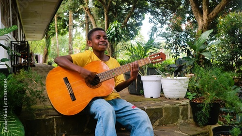 A stylish Indonesian teenager wearing a vibrant yellow t-shirt and blue jeans plays an acoustic guitar.