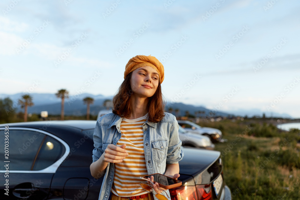 Fototapeta premium Young woman in an orange beanie relaxes beside a parked car on a sunny coastal road, holding a drink and enjoying a casual moment.