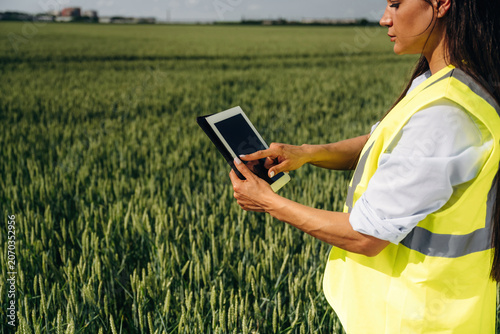 Female agricultural worker in reflective safety vest using tablet device in green wheat field under clear blue sky with distant buildings visible on the horizon