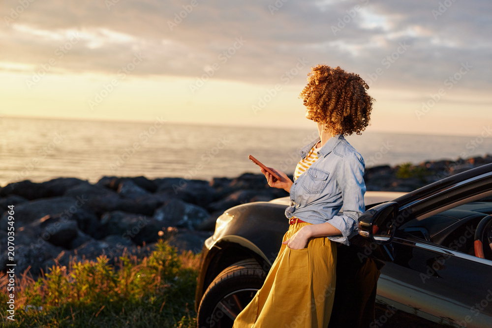 Fototapeta premium A woman with curly hair stands beside a car at the coast during sunset, looking at her phone. A candid lifestyle scene about travel, relaxation, and modern routines.