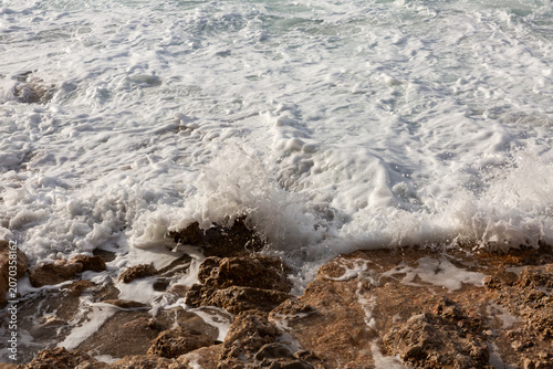 Bord de mer, gros plan sur une vague qui s’échoue et qui claque sur la roche brune en bord de plage, vue de face, écume de mer, mousse blanche