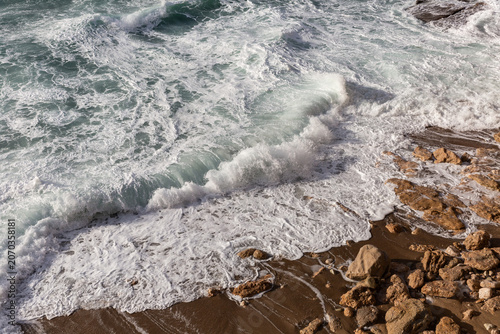 Rouleau d'une vague qui s’étale sur le sable et la roche brune, bord de mer agité, gros plan, mousse blanche