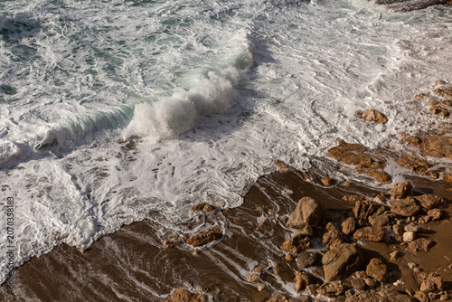 Bord de mer, gros plan sur une vague qui s’échoue sur le sable et la roche brune, mer agitée et vivante, écume de mer blanche