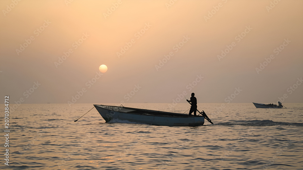 Fototapeta premium A lone fisherman steers his small boat across the calm sea at sunset, with another boat in the distance.