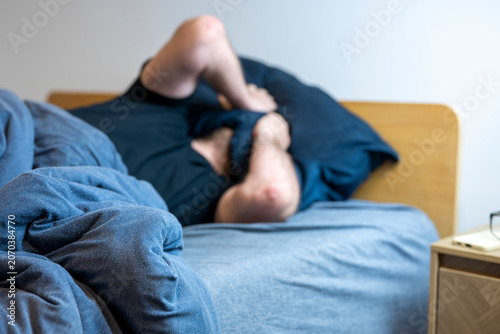 Obraz na plátně Man lies tangled in blue bedding, knees drawn to chest