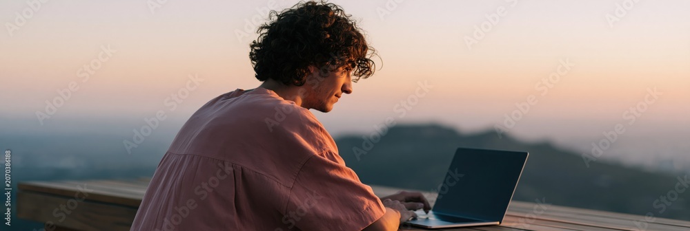 Obraz premium Young man working on laptop at sunset on a mountain balcony for remote work background and productivity wallpaper
