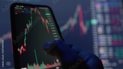 A dedicated trader intently focuses on a mobile phone that shows various stock charts, intricately revealing fluctuations in market trends illuminated by vibrant and colorful digital displays