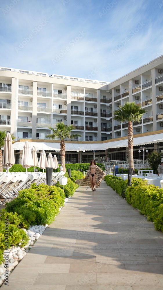 Fototapeta premium Tropical Resort Escape: A woman in a flowing cover-up walks down a scenic path towards a luxurious beachfront resort, palm trees swaying gently under a clear blue sky.