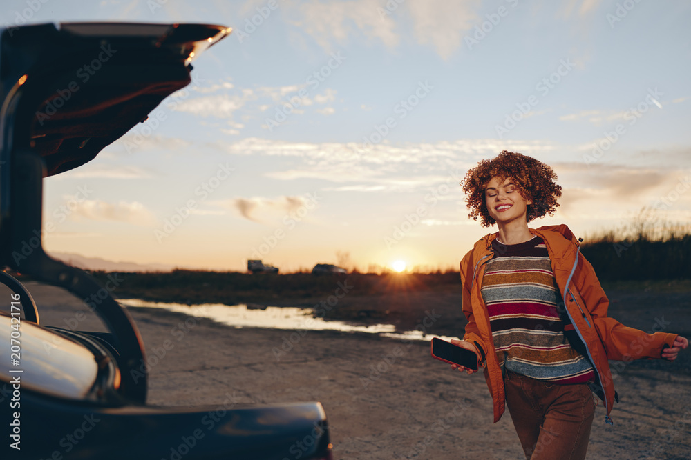 Fototapeta premium Smiling woman with curly hair by a car on a seaside road at sunset, travel lifestyle, warm sky, casual outfit, outdoor moment, soft natural light