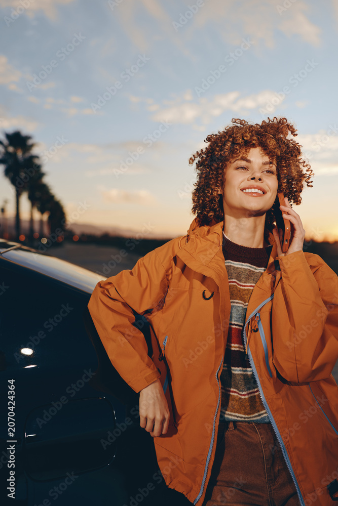 Fototapeta premium Woman outdoors, smiling at sunset, curly hair in warm light, wearing orange jacket and casual sweater, confident lifestyle travel mood, roadside background, soft evening breeze
