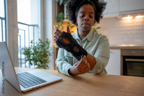 Φωτογραφία Focused woman adjusting orthopedic wrist brace while sitting at table with laptop