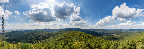 Oberotterbach shows its green hills and woodlands under a clear sky with clouds floating above.