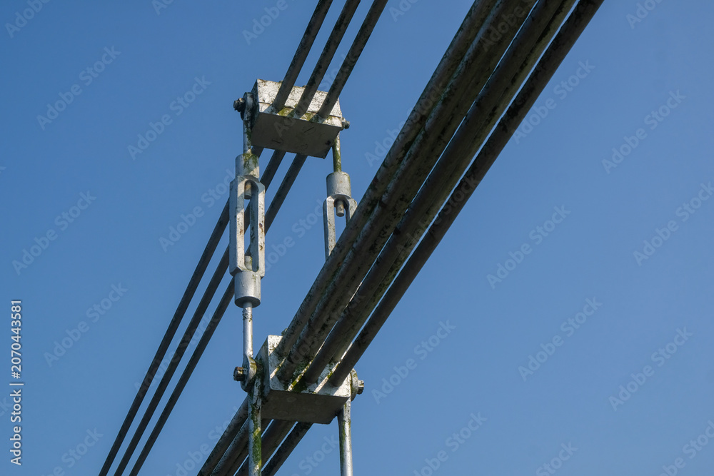 Fototapeta premium Close-up of steel cable tensioner and turnbuckle on a suspension bridge against clear blue sky. Structural engineering detail of bridge cable system. Industrial infrastructure and architecture concept