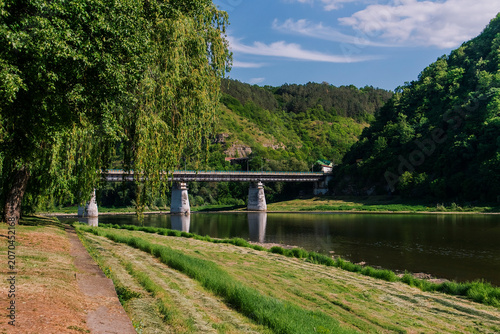 Peaceful riverside park in Zalishchyky, Ukraine, featuring graceful willow trees, a bridge spanning the Dniester River, and lush green hills.