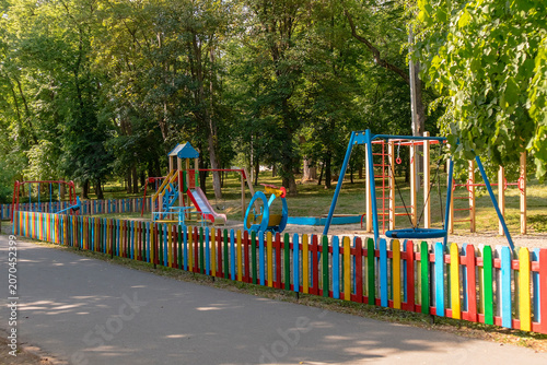 Bright children's playground enclosed by a colorful wooden fence, with swings, slide, and climbing equipment set among lush green trees in a public park.