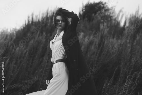 Monochrome fashion shot of a woman in a white dress and long coat standing in tall grass, dramatic pose, wind swept hair, outdoor moody portrait with strong contrast