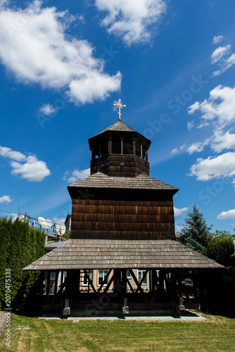 Traditional wooden church bell tower in Chortkiv, Ternopil region, Ukraine, standing beneath a bright blue sky with scattered clouds.