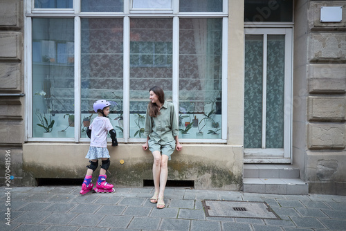 Mother watches daughter in roller skates and protective gear on sidewalk