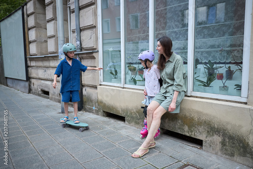 Mother watches children skateboarding and rollerblading on a city sidewalk