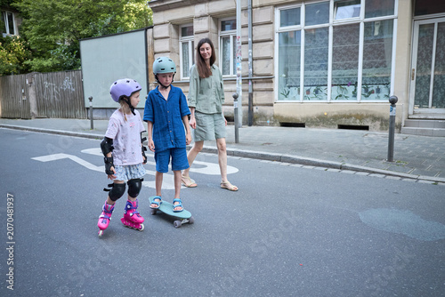 Mother watches children rollerblading and skateboarding on city street