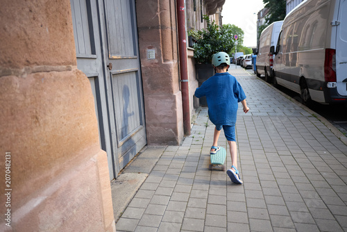 Young boy in blue outfit rides a skateboard on a paved sidewalk wearing a helmet