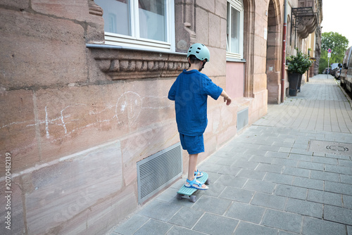 Young boy in blue outfit and helmet rides a skateboard down a city sidewalk
