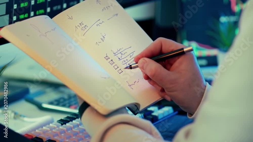 Writing tasks in a planner with pen while analyzing financial market trends on computer screen in a home office setup
