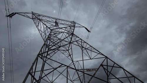 A dramatic, low-angle shot of a power pylon towering against dark overcast clouds. The moody lighting evokes an ominous energy crisis, framing the stark industrial latticework as symbol of grid strain