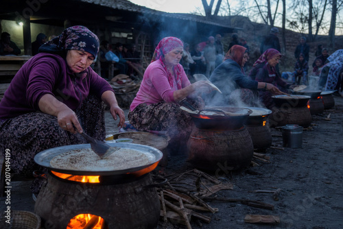 Women wearing traditional headscarves cook food in large metal pans over open wood fires at an outdoor communal gathering.