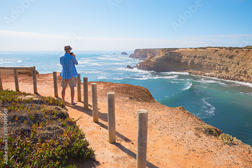 Картината върху платно vacationer stands near a cliff to take photos of  Algarve's steep coastline with a mobile phone