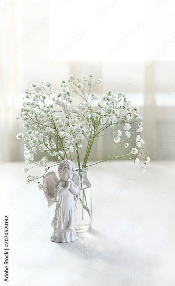 Naklejka premium Beautiful angel baby figurine and bouquet of white gypsophila flowers in glass vase on table, abstract light background. symbol of faith, pray, believe, christianity. romantic, lovely atmosphere
