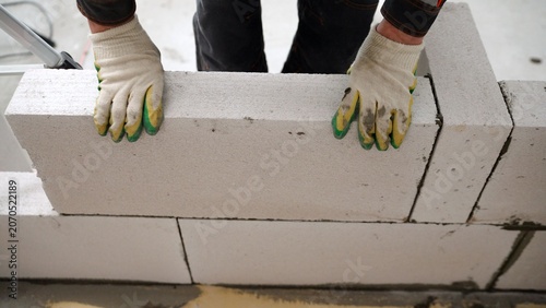 Construction worker in gloves placing a large aerated concrete block while building a wall at a construction site.