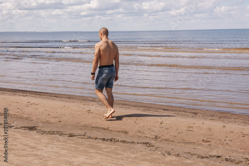 Shirtless man walking barefoot along a sandy beach by the sea on a sunny summer day. Rear view of an active adult enjoying a peaceful seaside walk during vacation.