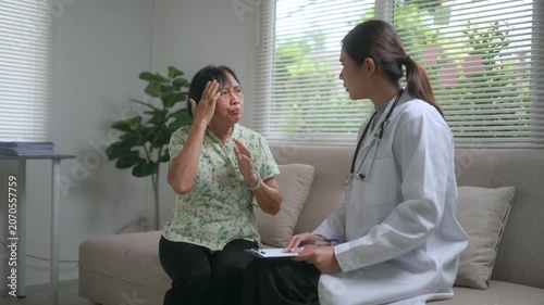 Senior asian woman sitting on a sofa describing her severe headache and migraine symptoms to a female doctor during a home visit, while the physician takes notes on a clipboard
