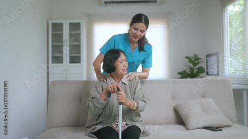 Young female nurse in scrubs giving a relaxing shoulder massage to an elderly woman sitting on a sofa with a walking stick, providing support and care in a cozy home living room