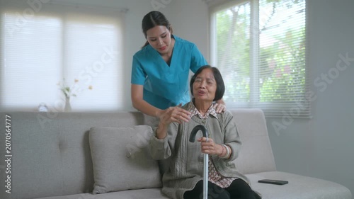 Kind young female nurse massaging the shoulders of a senior patient with a walking cane, providing home care service, physical therapy, and support in a comfortable living room setting