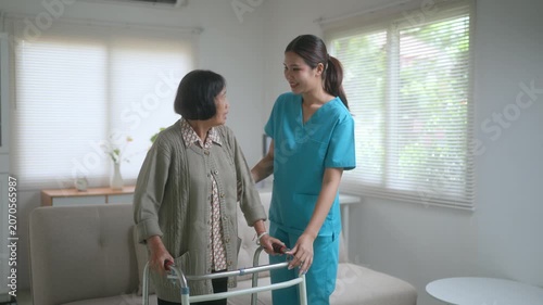 Young Asian female nurse assisting an elderly woman with a walker during a home visit, providing physical therapy and support for rehabilitation in a bright and comfortable living room