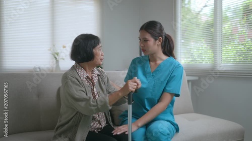 Cheerful young asian caregiver talking and supporting a smiling senior woman with a walking stick, providing assistance and companionship during a home visit in the living room