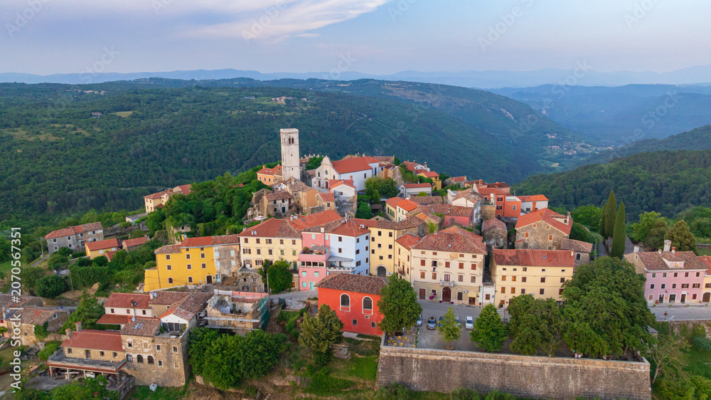 Fototapeta premium Drone captures medieval hilltop town of Oprtalj located in inland Istria. Historic stone houses, cypress trees, and ancient bell tower stand above picturesque countryside vineyards and fields.