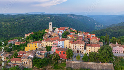 Drone captures medieval hilltop town of Oprtalj located in inland Istria. Historic stone houses, cypress trees, and ancient bell tower stand above picturesque countryside vineyards and fields.