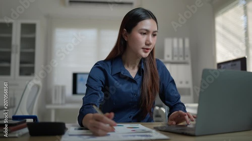 Professional asian woman analyzing financial charts on documents while using a laptop computer at her desk in a modern office, focused on business growth and market analysis