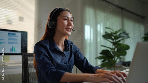 Young Asian woman wearing a headset working as a customer service representative, talking and smiling while looking at a laptop computer screen in her home office during the evening
