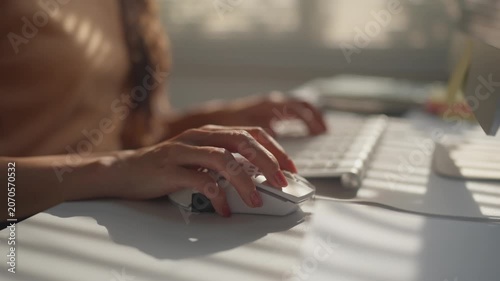 Woman's manicured hands working on a desktop computer, typing on the keyboard and using the mouse at a sunlit desk in a modern office, showcasing a focused and productive work environment