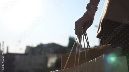 Female arm holds full paper packets walking along urban street. Hand of young woman carries shopping bags going on city alley after purchases. Concept of seasonal sales and discounts. Close up Slow mo