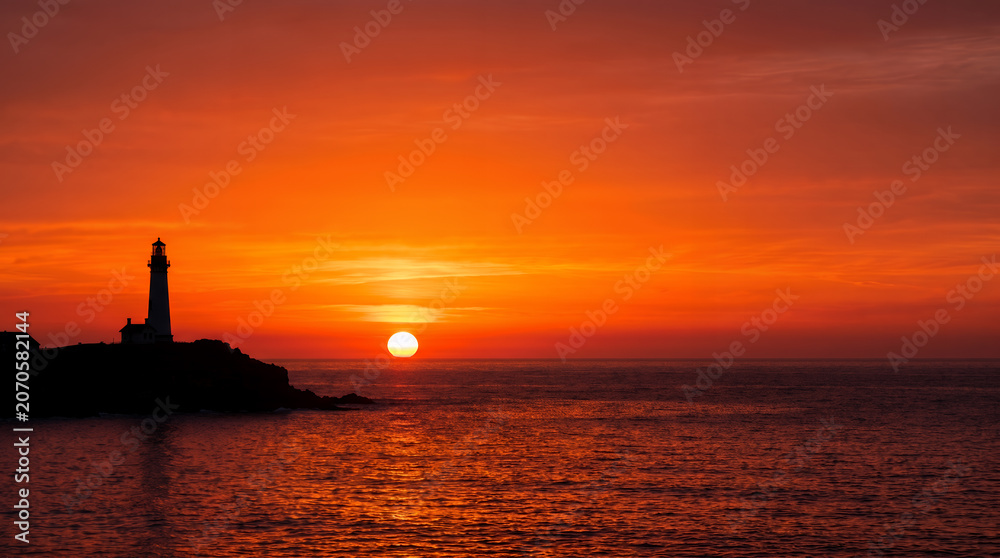 Fototapeta premium Silhouette of a coastal lighthouse on a rocky cliff during a vibrant fiery sunset over the ocean