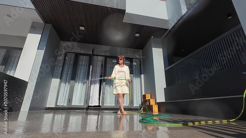 Woman in yellow outfit does concrete cleaning with green hose on modern patio. Strong water spray washes dusty tiles, keeping beautiful townhouse yard neat and perfectly maintained.