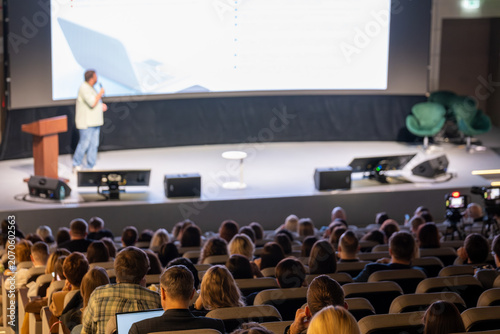 Presenter on stage delivering business presentation to large audience in modern conference auditorium, attendees listening and taking notes.