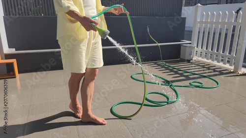 Barefoot woman does patio cleanup with green hose on sunny terrace. Water stream washes outdoor tiles, while white fence on background creates neat and peaceful house backyard atmosphere.